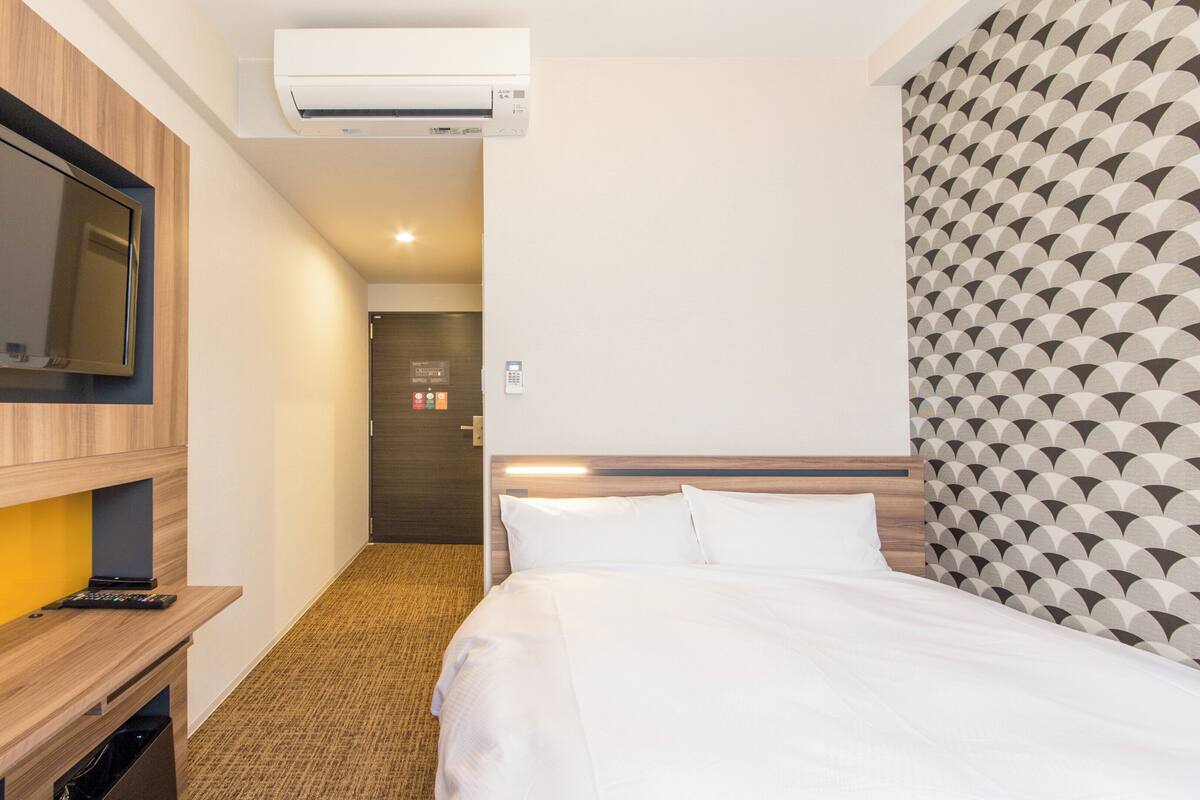 A hotel room with a bed, a TV mounted on the wall, a desk with a remote control, and an air conditioning unit.