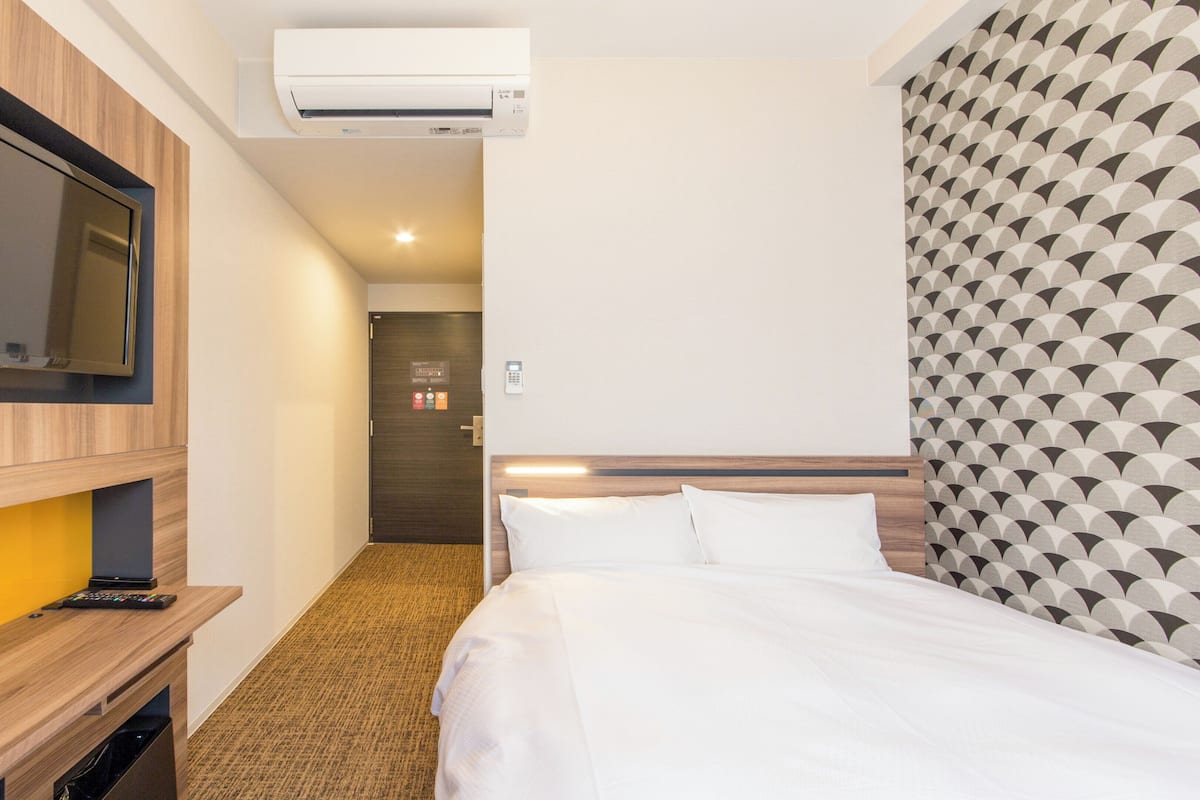 A hotel room with a bed, a TV mounted on the wall, a desk with a remote control, and an air conditioning unit.