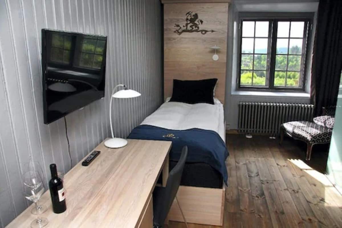 Une chambre avec un lit, un bureau sur lequel se trouve une bouteille de vin, une chaise, un téléviseur fixé au mur et une fenêtre donnant sur un beau paysage.
