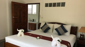 Una cama bien hecha con cabecera de madera, ropa de cama blanca y almohadones decorativos. Se ve una puerta de madera y una mesita con una lámpara en la habitación.