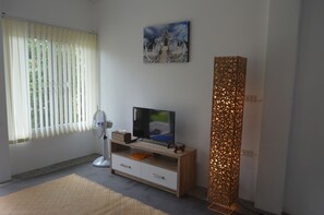 Ein Zimmer mit Fernseher auf einem Holzständer, einem Standventilator, einer gewebten Matte auf dem Boden und einer dekorativen Leuchte.