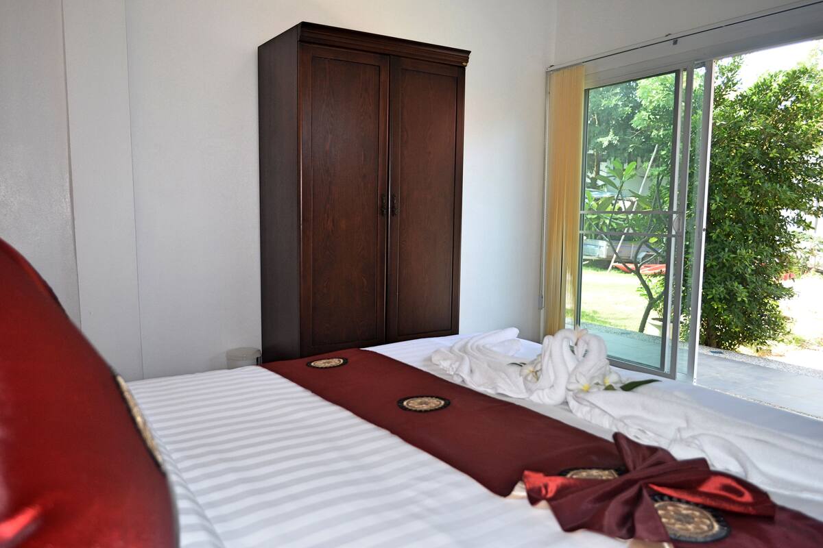 A hotel room with a bed, a wooden wardrobe, and a view of greenery through a glass door.