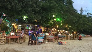 Un área de comedor al aire libre con mesas y sillas, rodeada de árboles y decorada con luces.