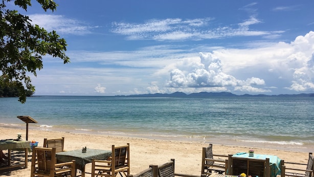 Un restaurante frente a la playa con mobiliario de madera y vistas al mar y a las montañas.