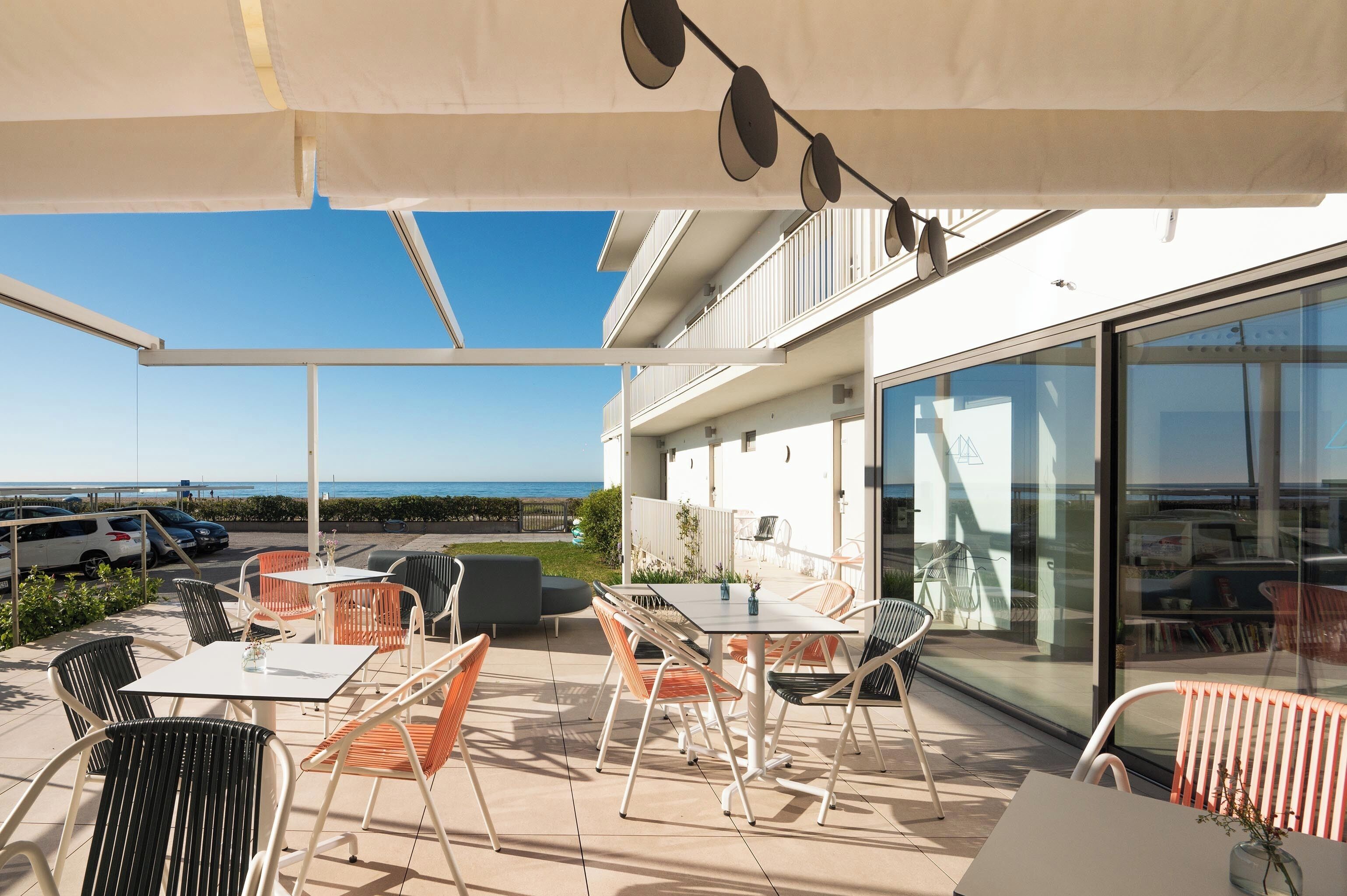 Un patio al aire libre con mesas y sillas, vista al mar y un edificio con fachada de vidrio.