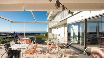 Un patio al aire libre con mesas y sillas, vista al mar y un edificio con fachada de vidrio.