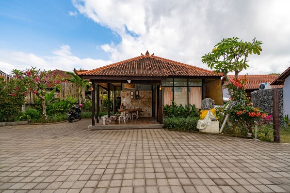 Une maison traditionnelle avec un toit en tuiles, un patio couvert et un jardin agrémenté de plantes et d’une statue.