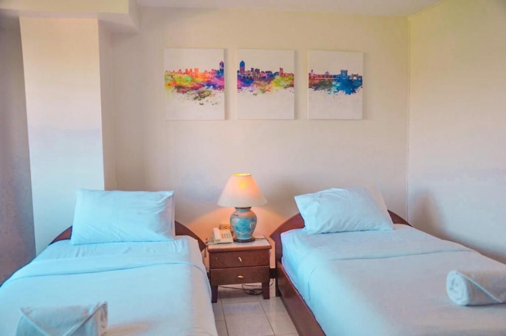 A hotel room with two beds, a nightstand with a lamp, and three cityscape paintings on the wall.