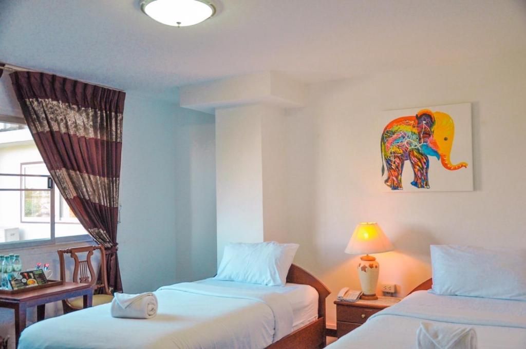 A hotel room with two beds, a window with curtains, a desk with a lamp, and a colorful elephant painting on the wall.