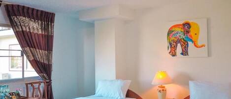 A hotel room with two beds, a window with curtains, a desk with a lamp, and a colorful elephant painting on the wall.