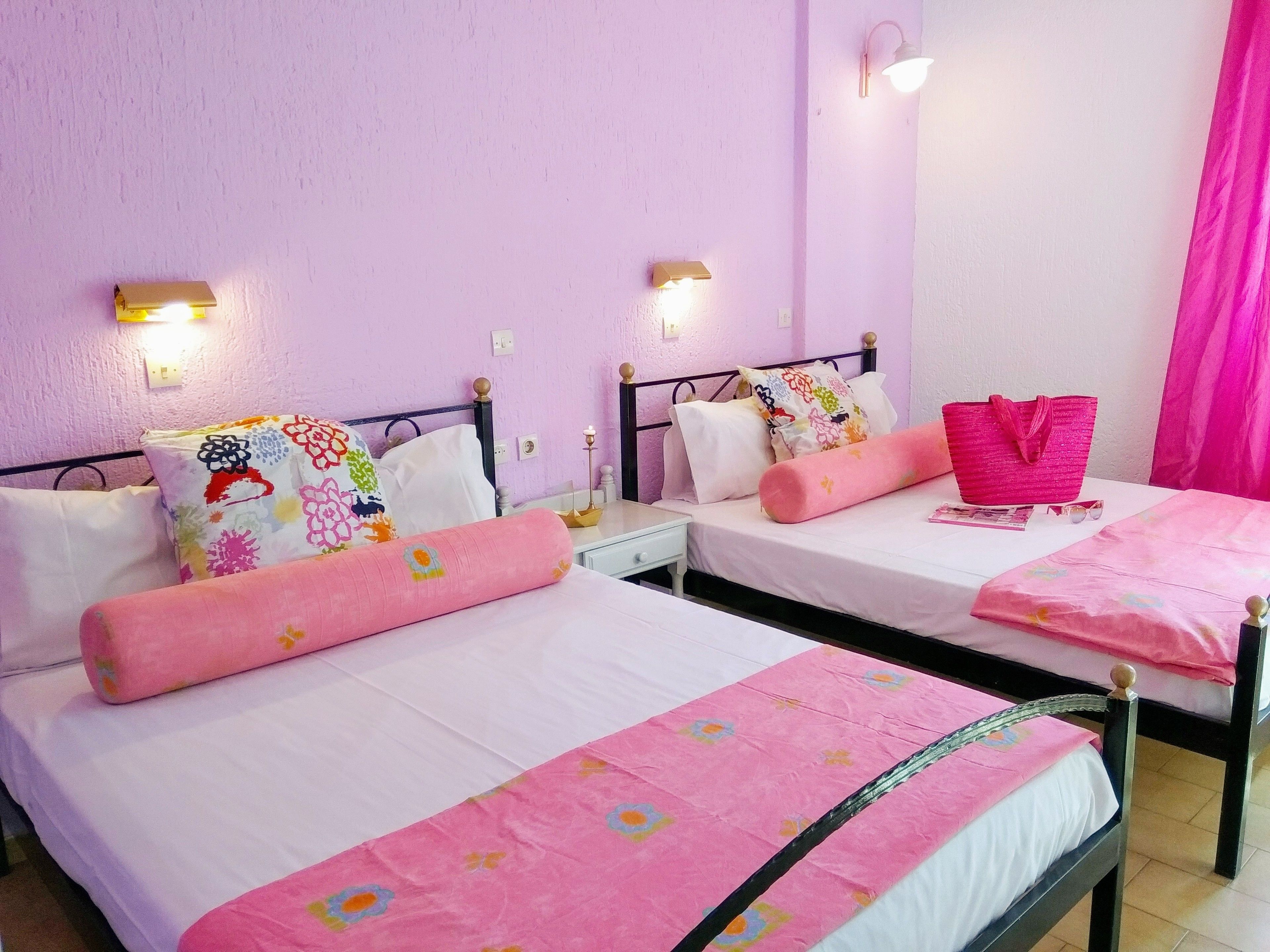 Una camera con due letti, ognuno con una coperta rosa e cuscini bianchi. Sono presenti luci a parete e una tenda rosa.