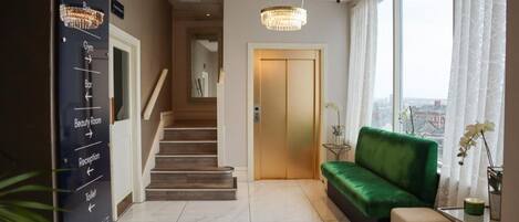 Un hall d’hôtel avec un escalier, un comptoir d’accueil, un canapé vert et une vue sur la ville grâce à de grandes fenêtres.