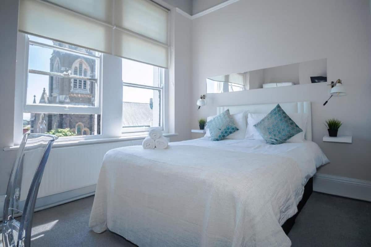 A bedroom with a large window, a bed with white bedding and patterned pillows, a bedside table with a lamp, and a small plant.