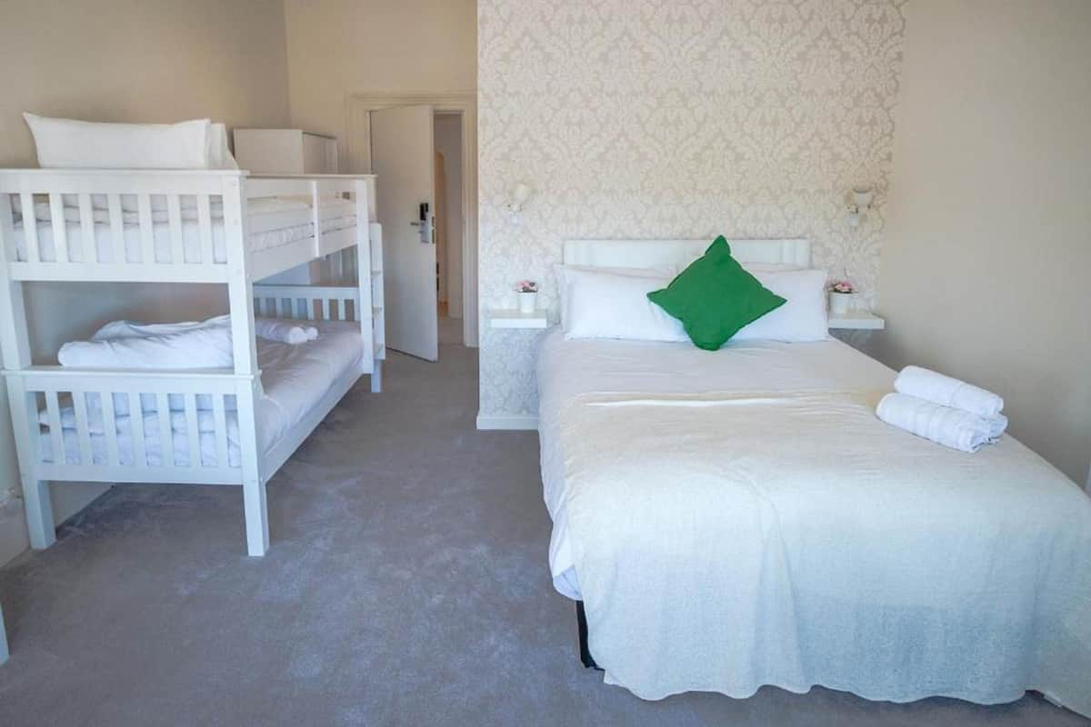 A hotel room with bunk beds, a single bed, white bedding, a green pillow, and a nightstand with a lamp.