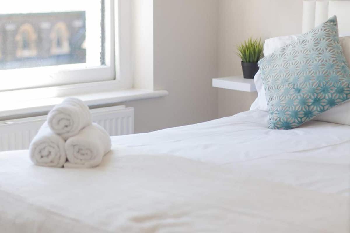 A neatly made bed with rolled white towels, a blue patterned pillow, and a small potted plant on a white bedside table.