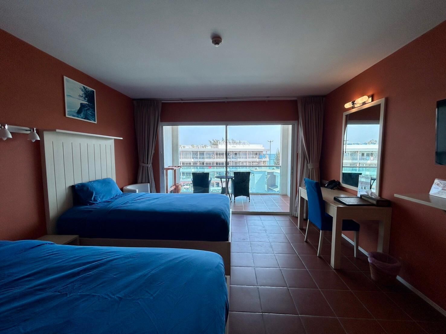 A hotel room with two beds, a desk, a chair, and a balcony with a view of buildings.