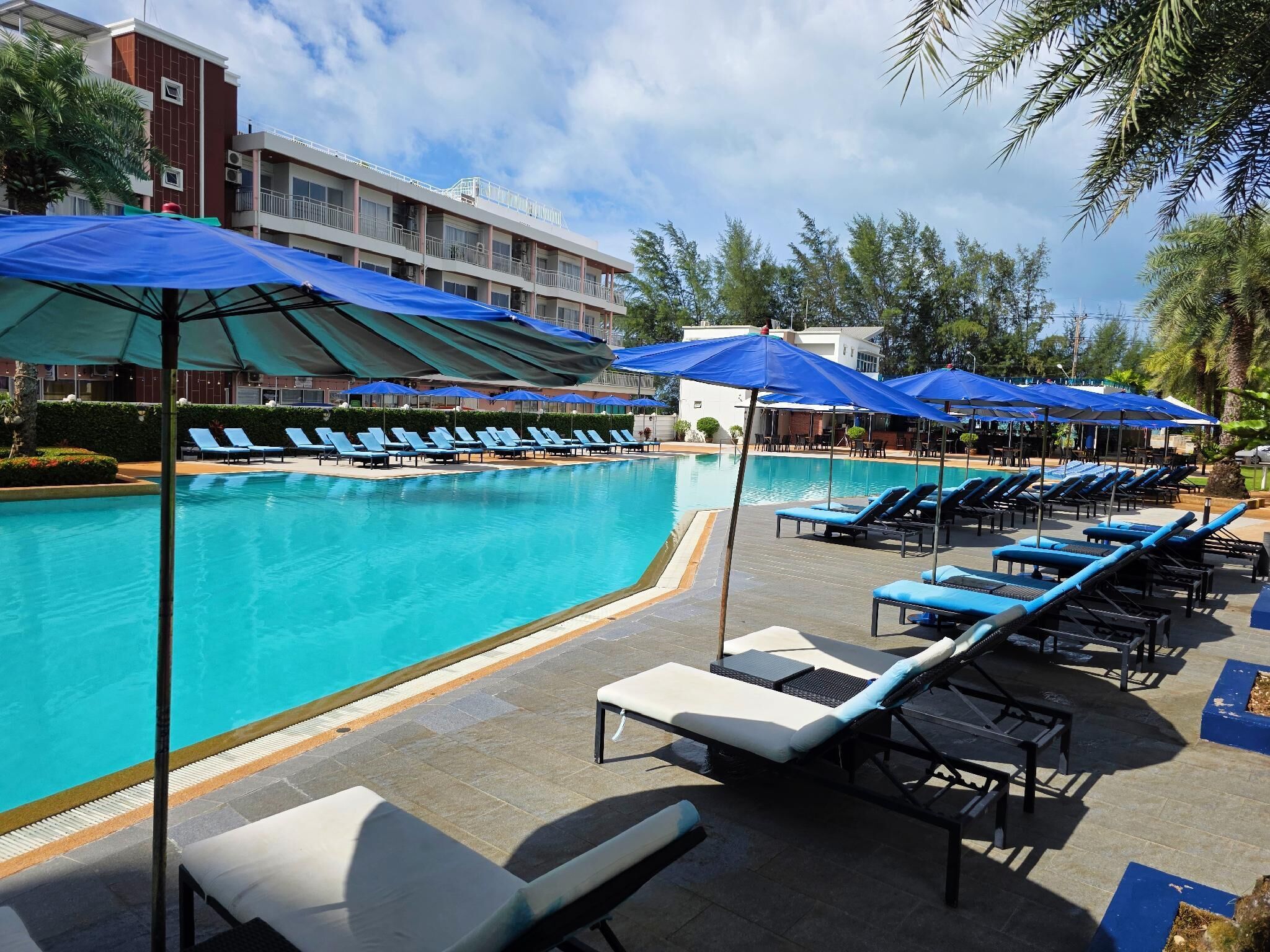 Un área junto a la piscina con sillones de descanso y sombrillas, una piscina azul cristalina y un edificio al fondo.