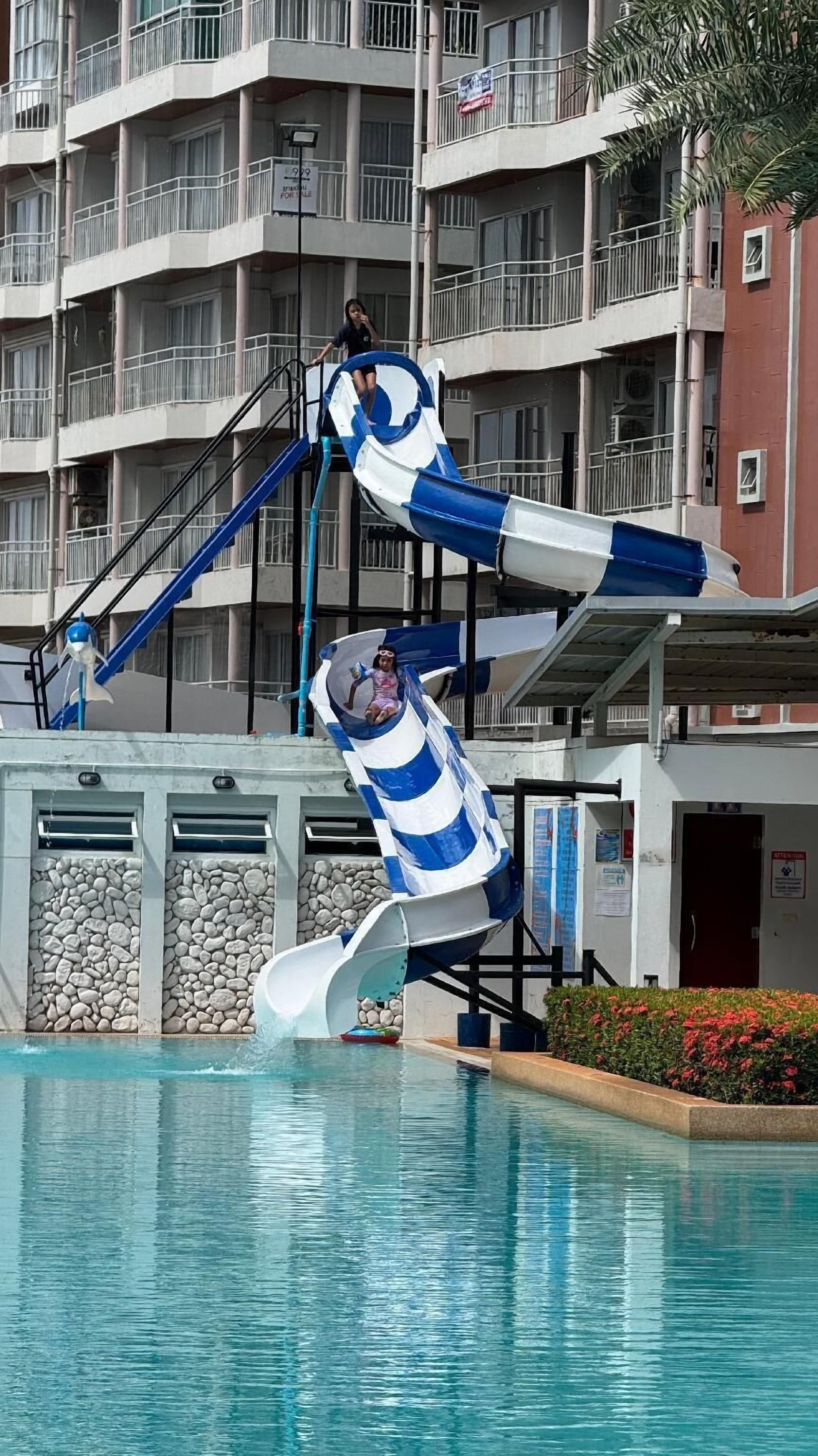A multi-story building with balconies, a pool area with a water slide, and a sign indicating a for sale property.
