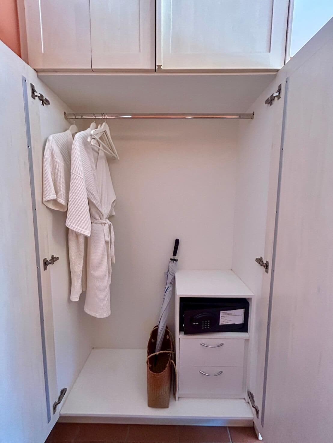 Ein kleiner, weißer Kleiderschrank mit zwei aufgehängten Bademänteln, eine kleine, weiße Kommode mit Schubladen und eine braune Tasche auf dem Boden.