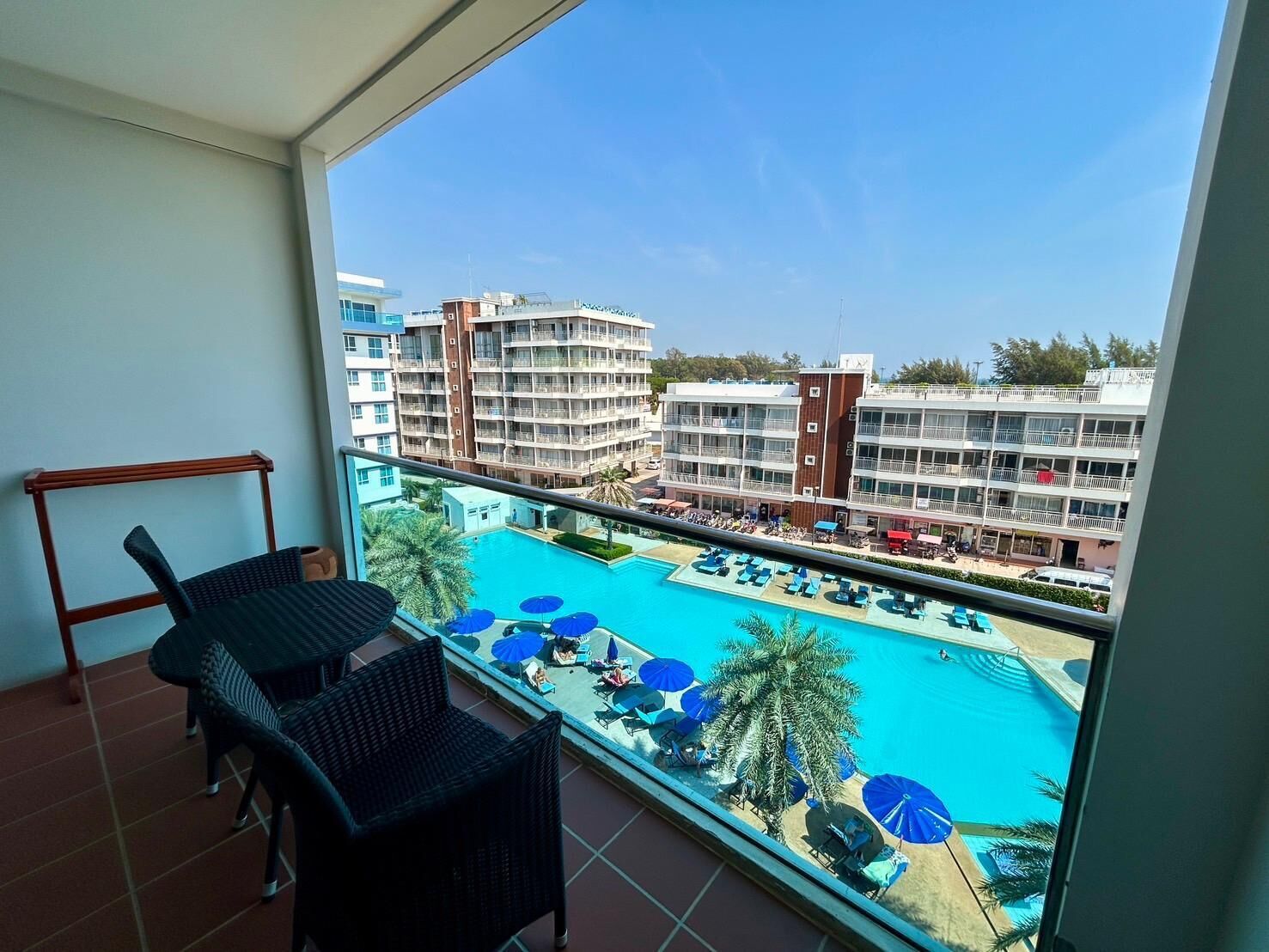 Un balcón con mobiliario de mimbre que da a una zona de piscina con sombrillas azules y palmeras.