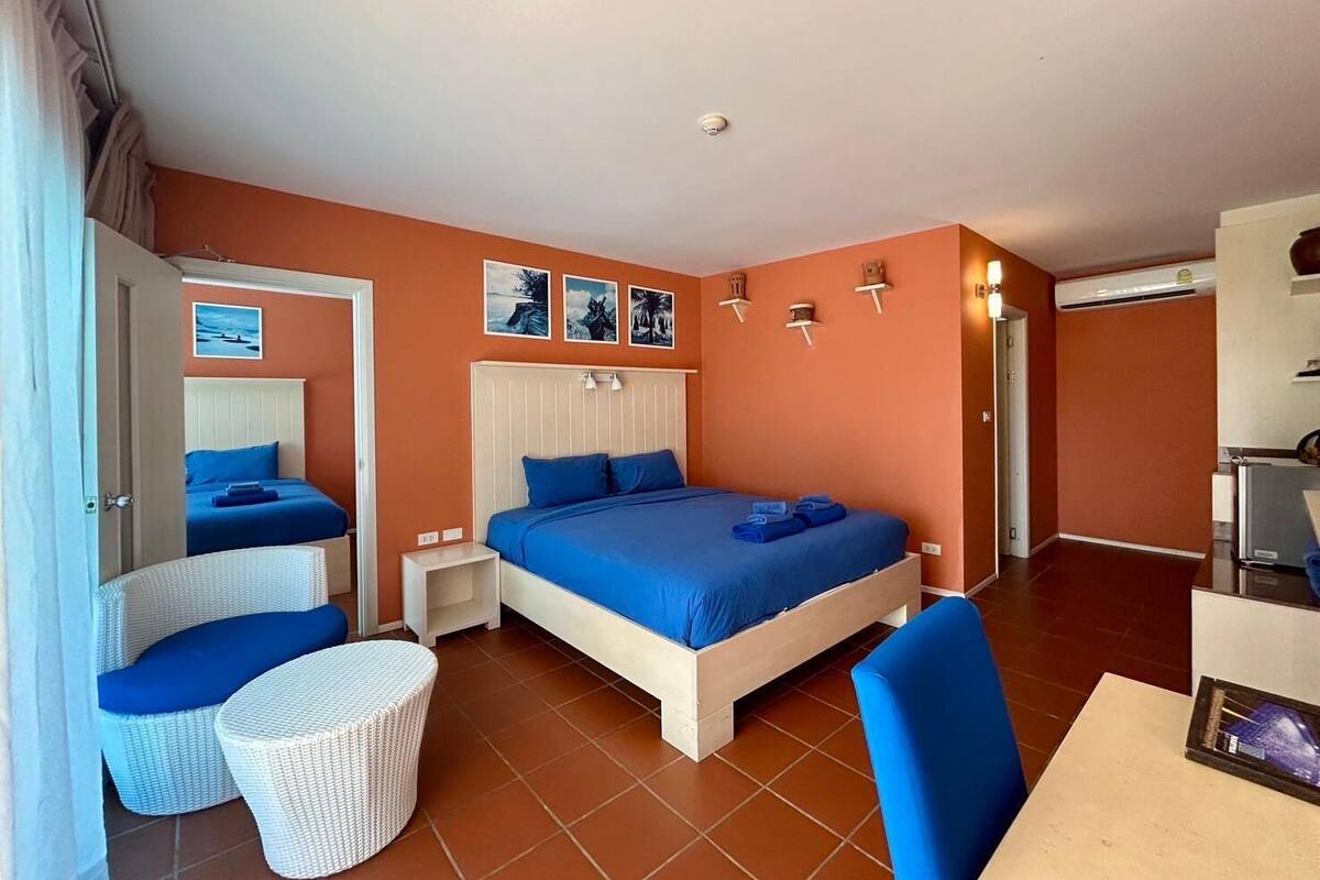 Quarto com cama grande, duas mesas de cabeceira, uma poltrona de vime, um banquinho pequeno e uma escrivaninha com cadeira.