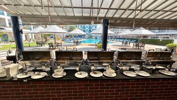 Montaje de bufé con varias estaciones de comida, asientos exteriores con sombrillas y una piscina al fondo.