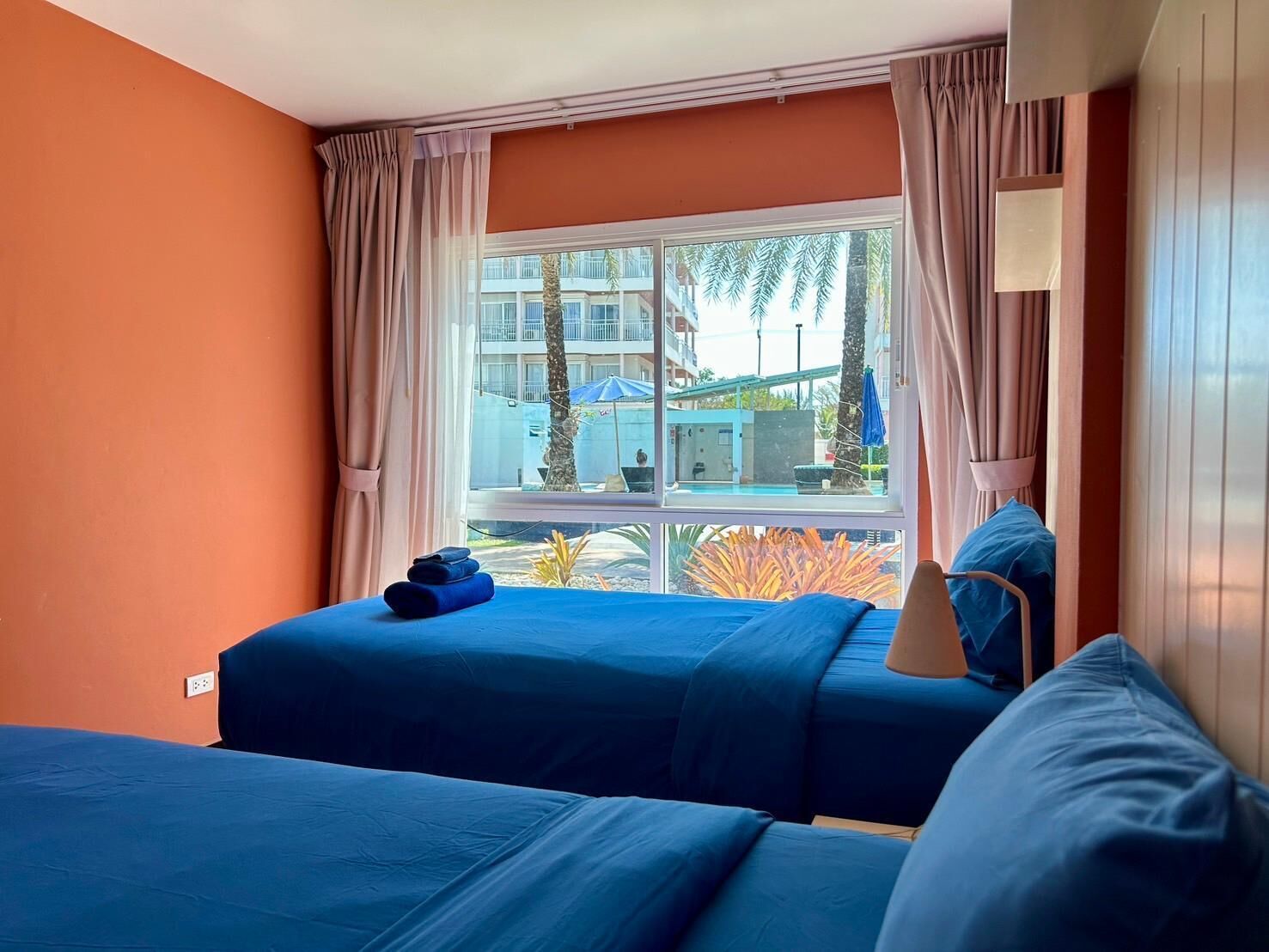 A hotel room with two beds, orange walls, a large window with curtains, and a view of a building and palm trees outside.