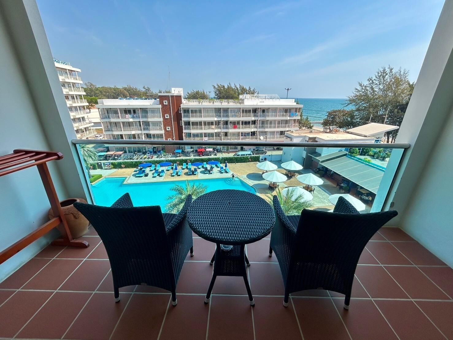 A balcony with two wicker chairs and a small table, overlooking a pool area with umbrellas and lounge chairs.