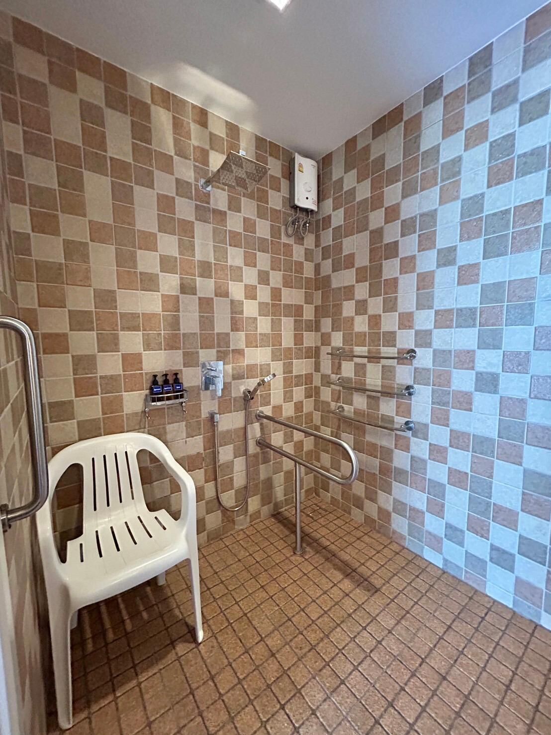 Un baño con pared de baldosas a cuadros, una silla de plástico blanca, un pasamanos y un panel de control de ducha.