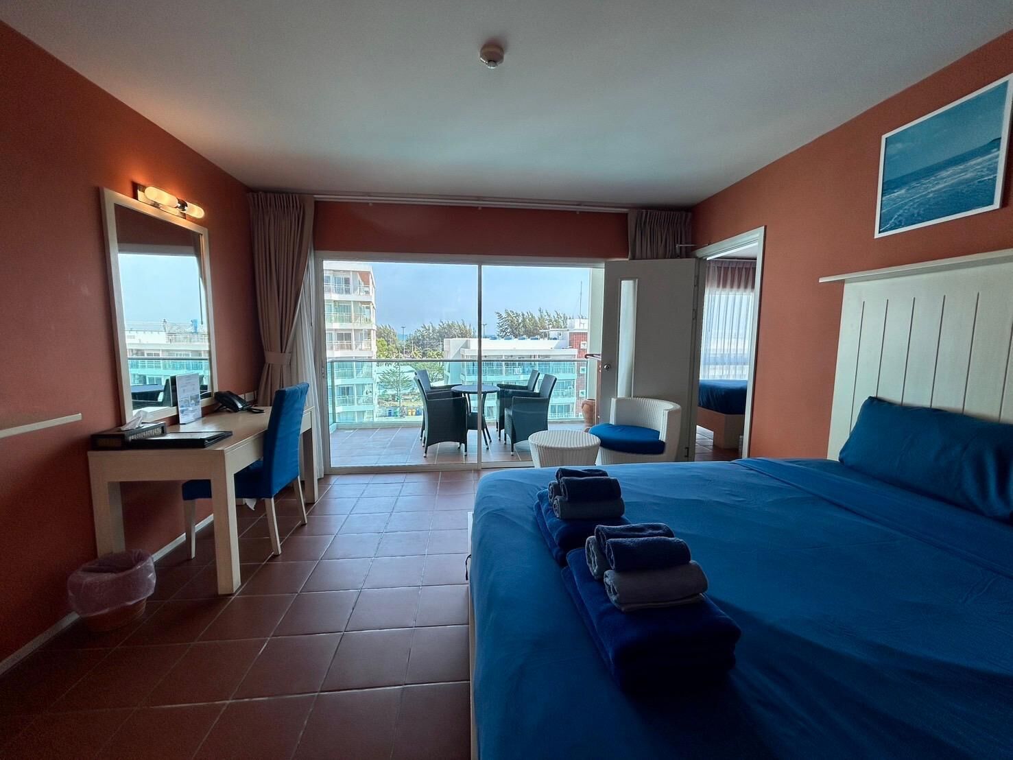 A hotel room with a bed, a desk, a chair, a small table, and a balcony with a view of buildings and trees.