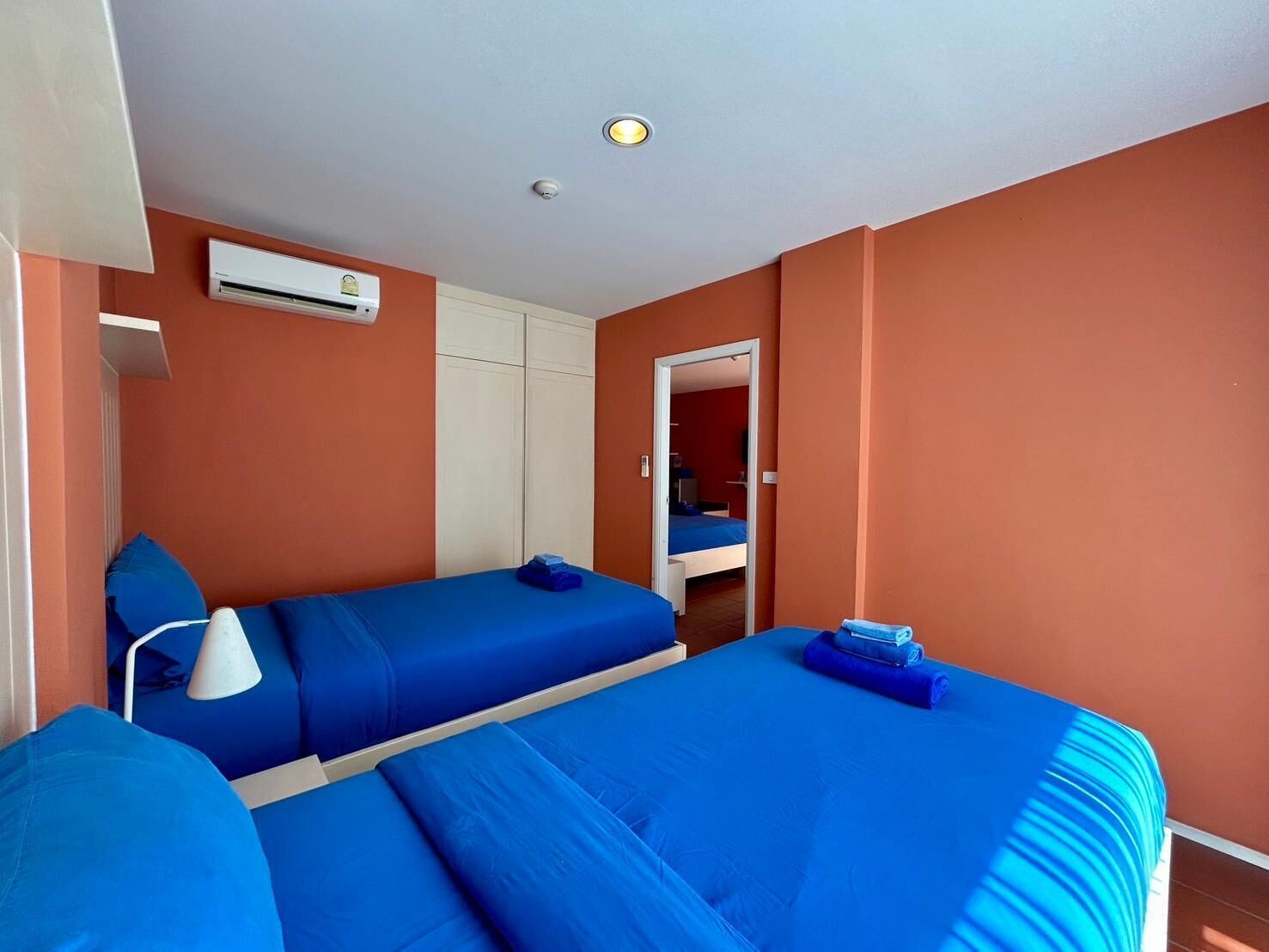 A room with two beds, a white air conditioning unit, a white lamp, and a door leading to another room.