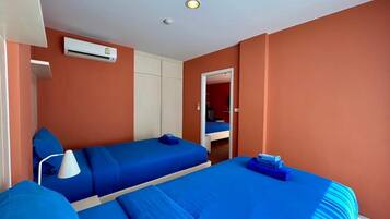 A room with two beds, a white air conditioning unit, a white lamp, and a door leading to another room.