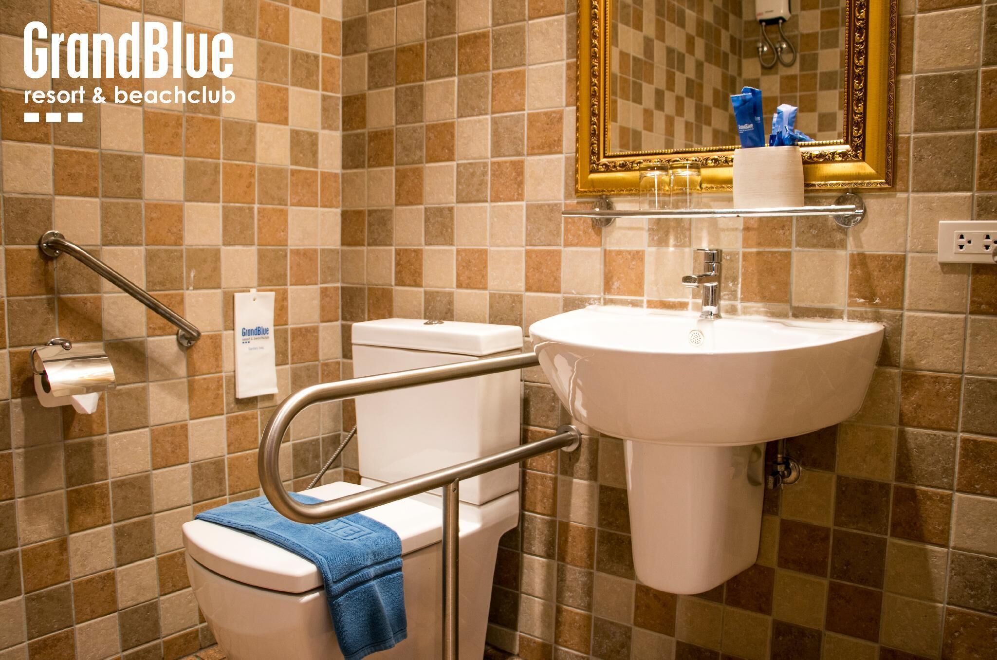 Un baño con inodoro, lavamanos y barras de apoyo, con pared de azulejos y espejo con marco dorado.