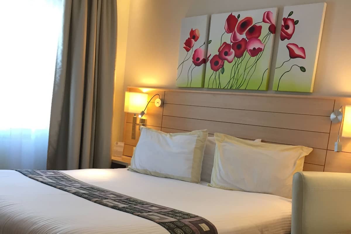 A hotel room with a large bed, a bedside table, a chair, and a painting of red flowers on the wall.