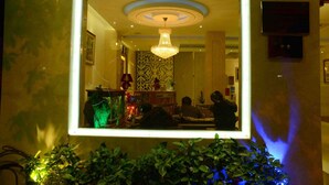 Un espacio interior bien iluminado con una araña, un espejo enmarcado que refleja la habitación y una planta en maceta al frente.