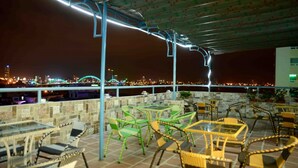Un patio al aire libre con mesas y sillas, vista de un paisaje urbano nocturno y una sección cubierta con techo estampado.