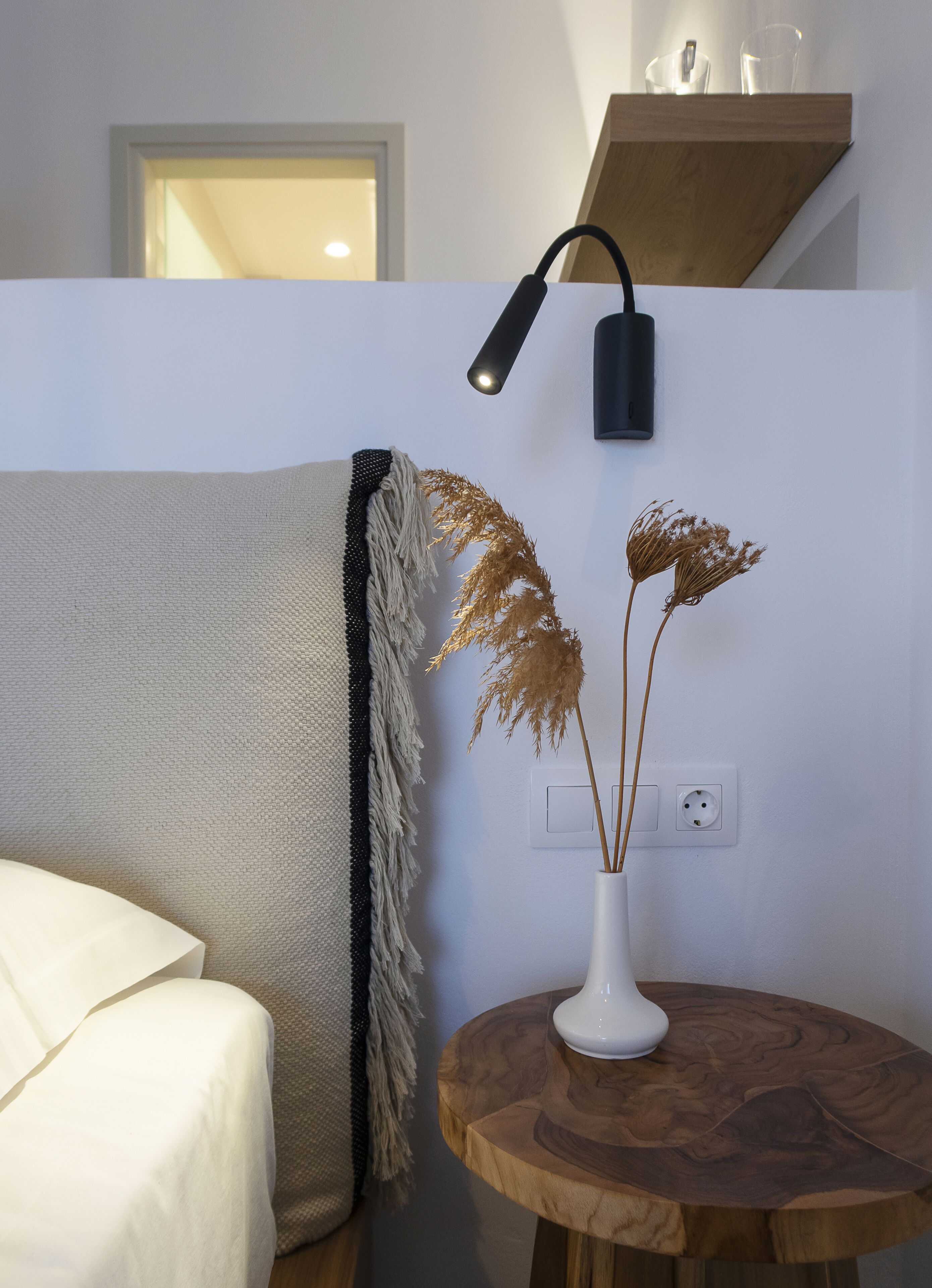 A room with a white bed, a round wooden side table with a white vase, and a wall-mounted lamp.