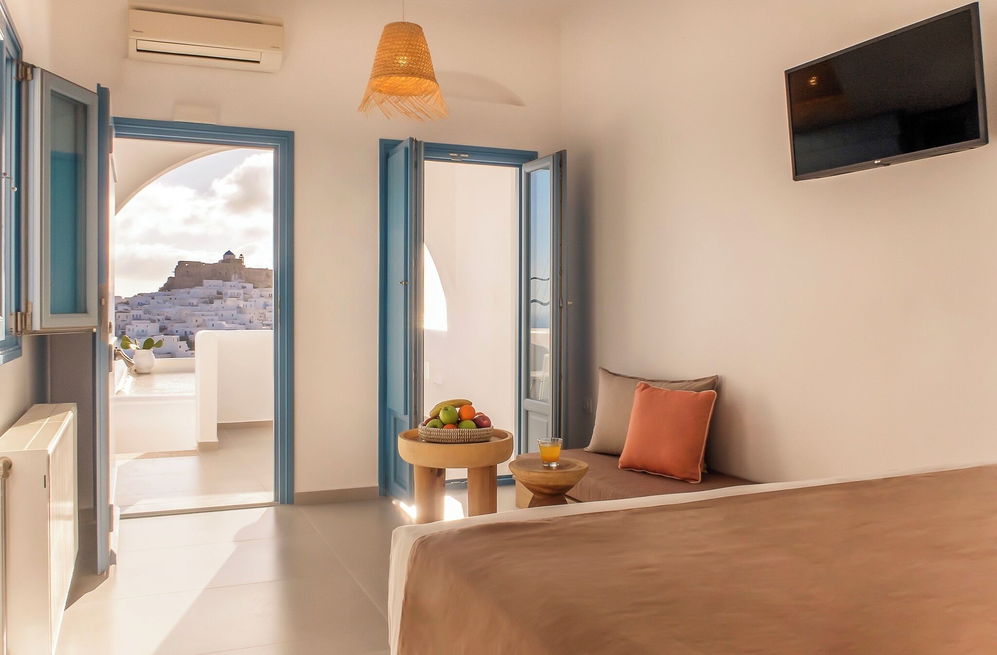 A hotel room with a bed, a TV, a fruit bowl, and a view of a white building.