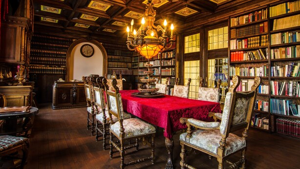 Una biblioteca con una mesa roja, estanterías de madera y una araña.
