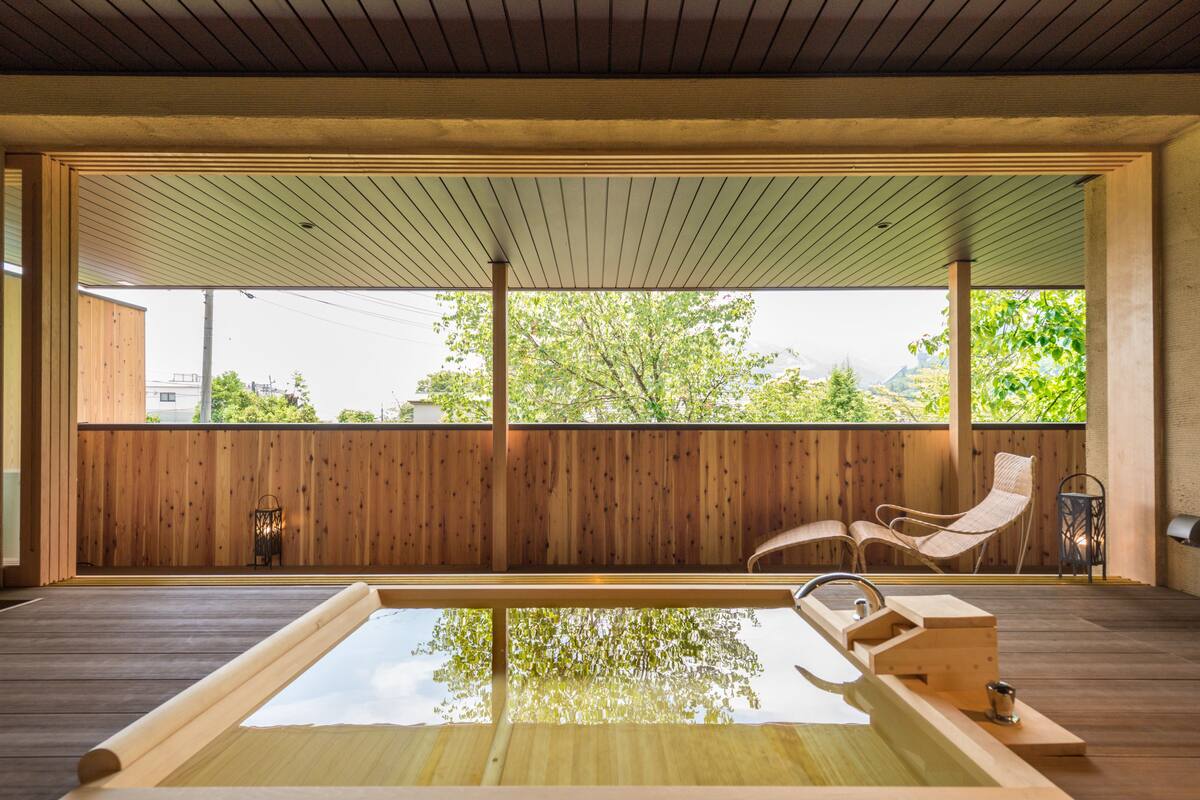 Une terrasse en bois avec un jacuzzi, deux fauteuils en osier et une vue sur les arbres et les bâtiments.