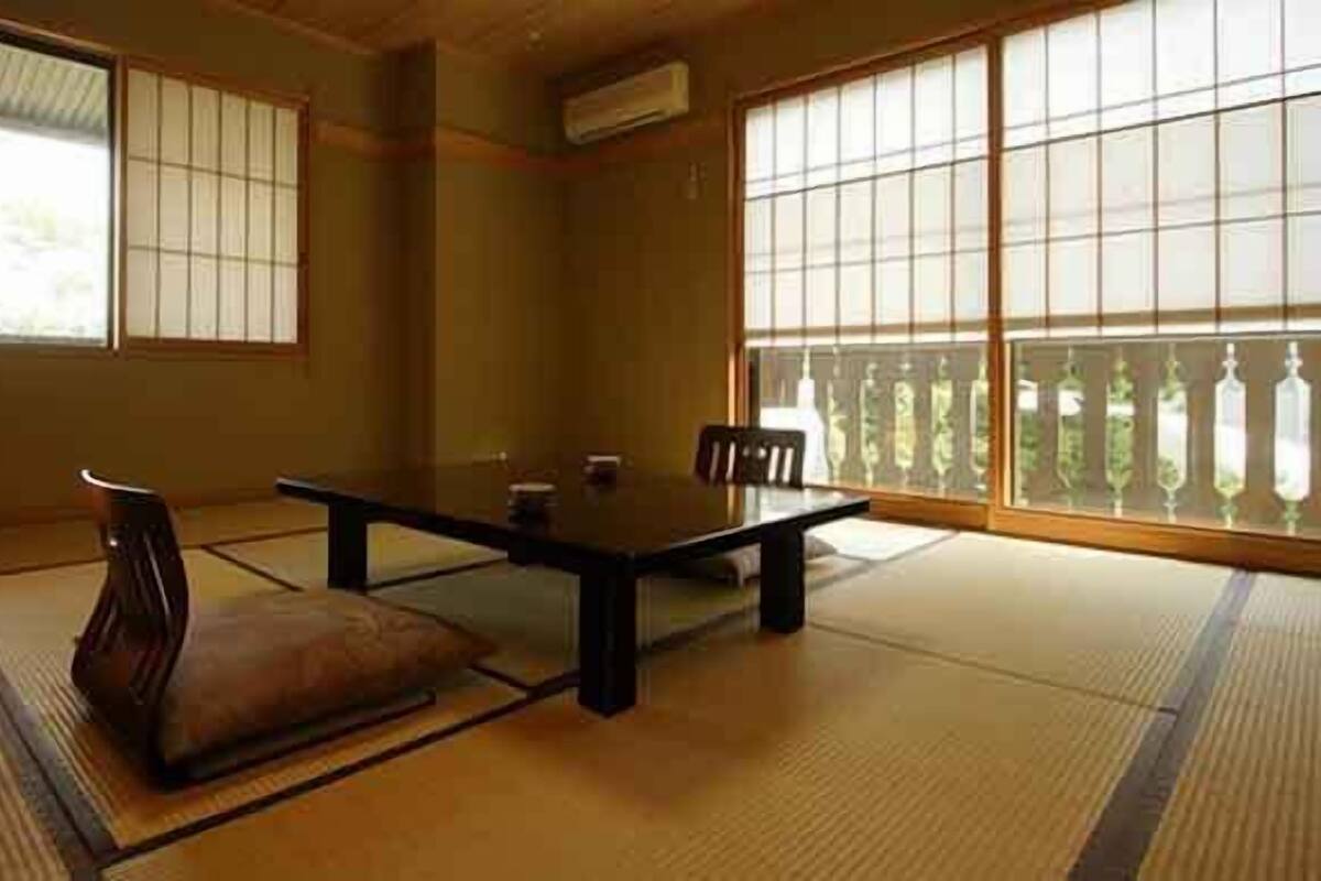 Une pièce avec un sol recouvert de tatamis, une petite table et des chaises, de grandes fenêtres et une esthétique japonaise traditionnelle.