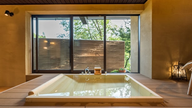 A modern spa room with a large wooden bathtub, a sliding glass door leading to a garden, and a small wooden stool. - Hotel Hakuba Hifumi (Hakuba)