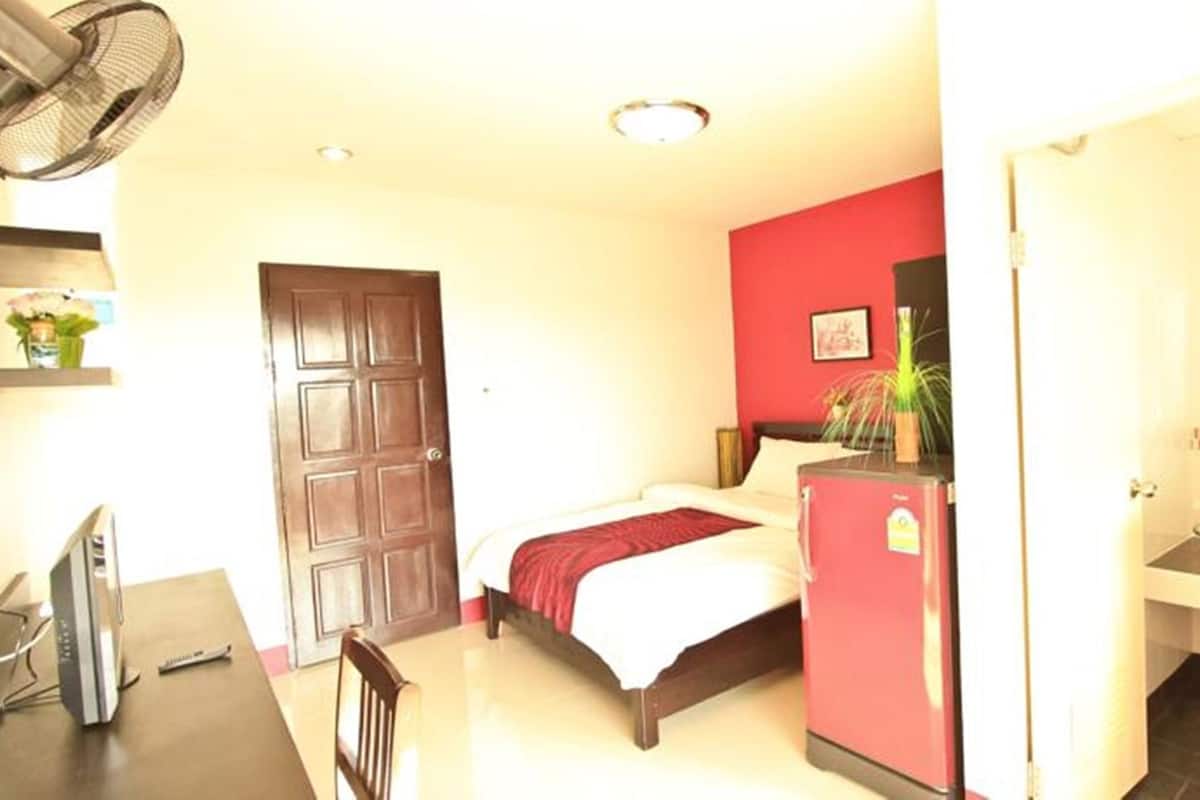 A hotel room with a bed, a desk with a chair, a small refrigerator, a sink, and a fan.