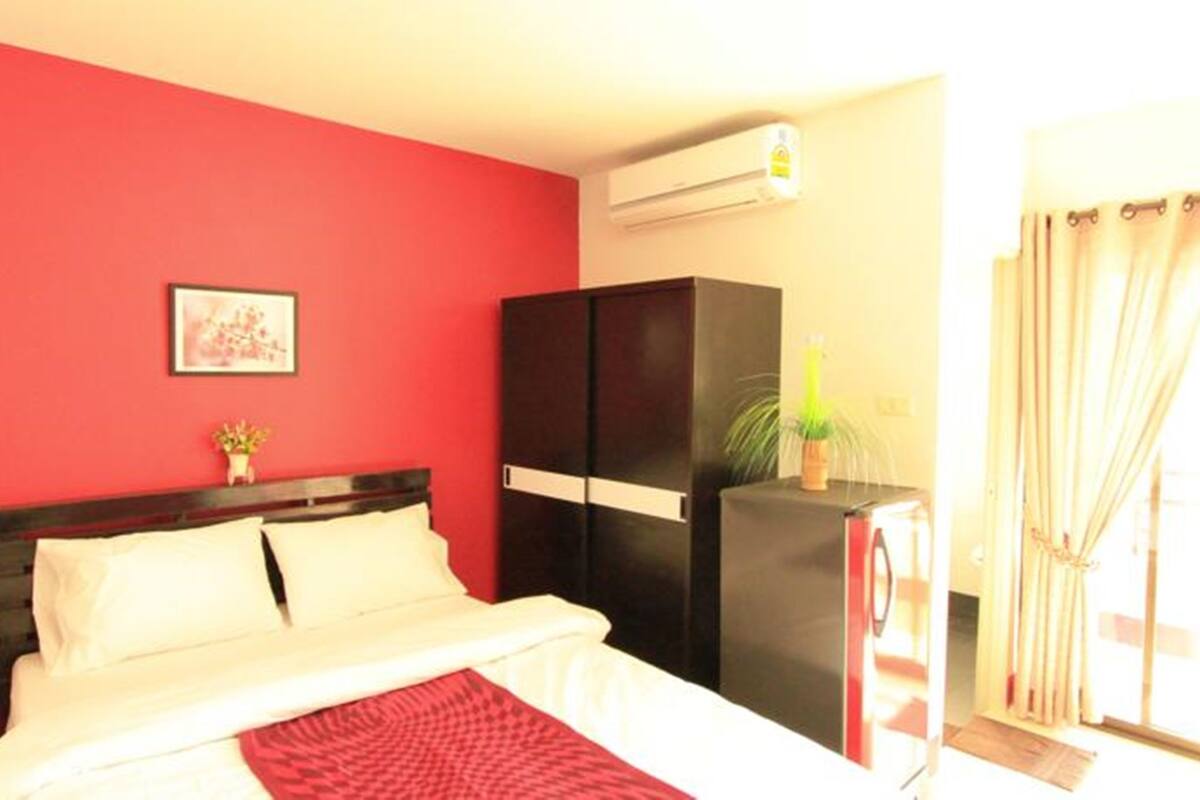 A hotel room with a red accent wall, a bed with white bedding, a black wardrobe, and a small potted plant.