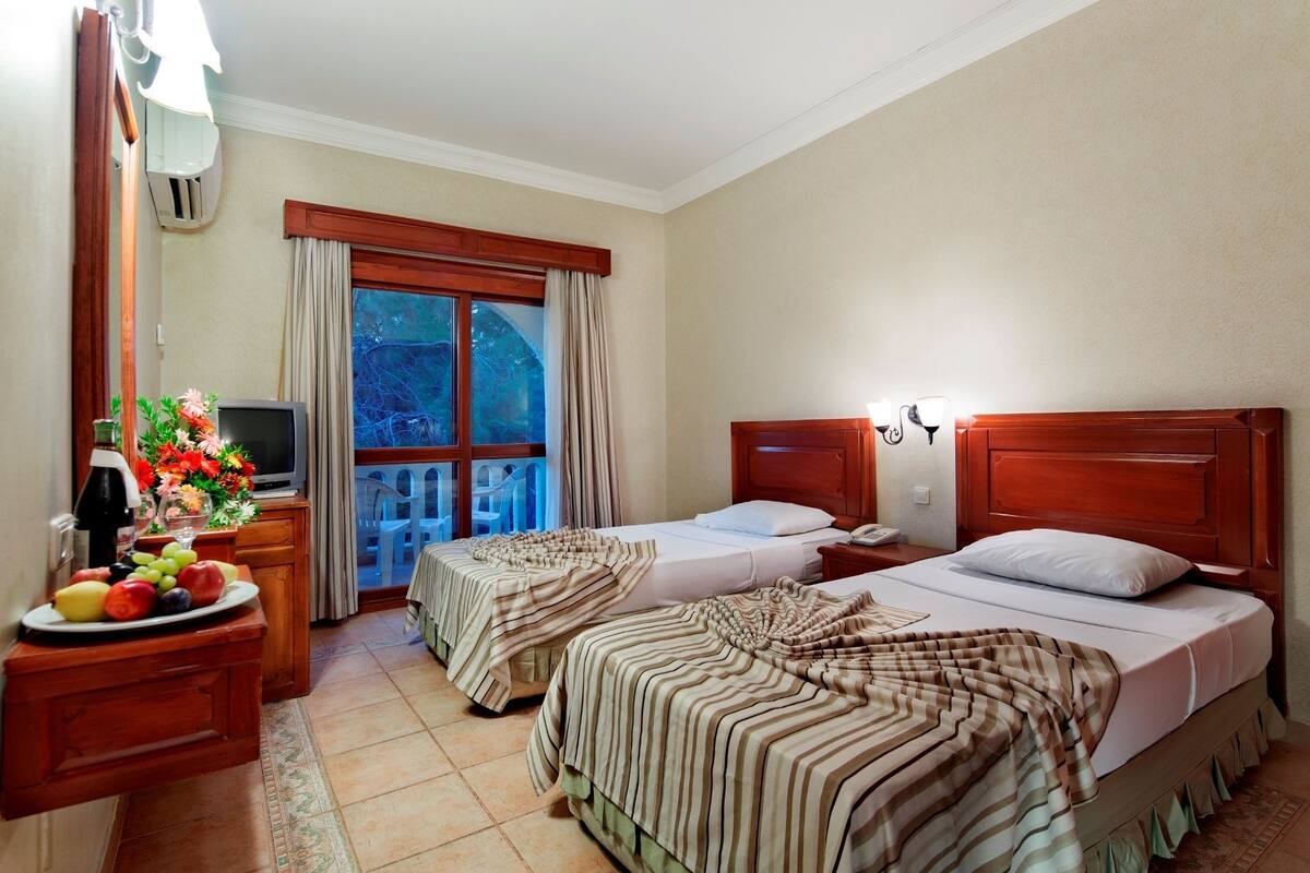Ein Hotelzimmer mit zwei Betten, einem Fernseher, einem kleinen Tisch mit Früchten und einem Balkon mit Vorhängen.