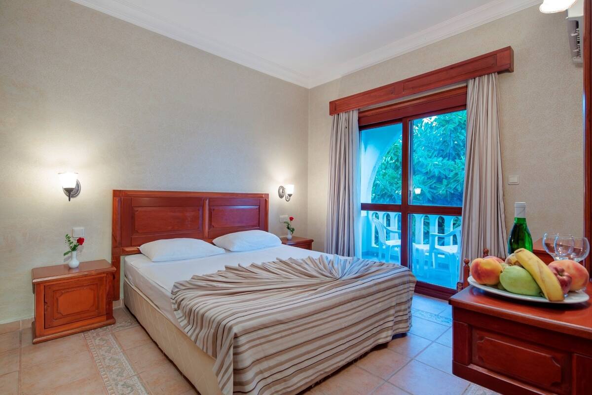 Ein Schlafzimmer mit einem großen Bett, einem Holz-Kopfteil, zwei Nachttischen mit Lampen, einem Beistelltisch mit einer Vase und Blick ins Freie durch ein großes Fenster.