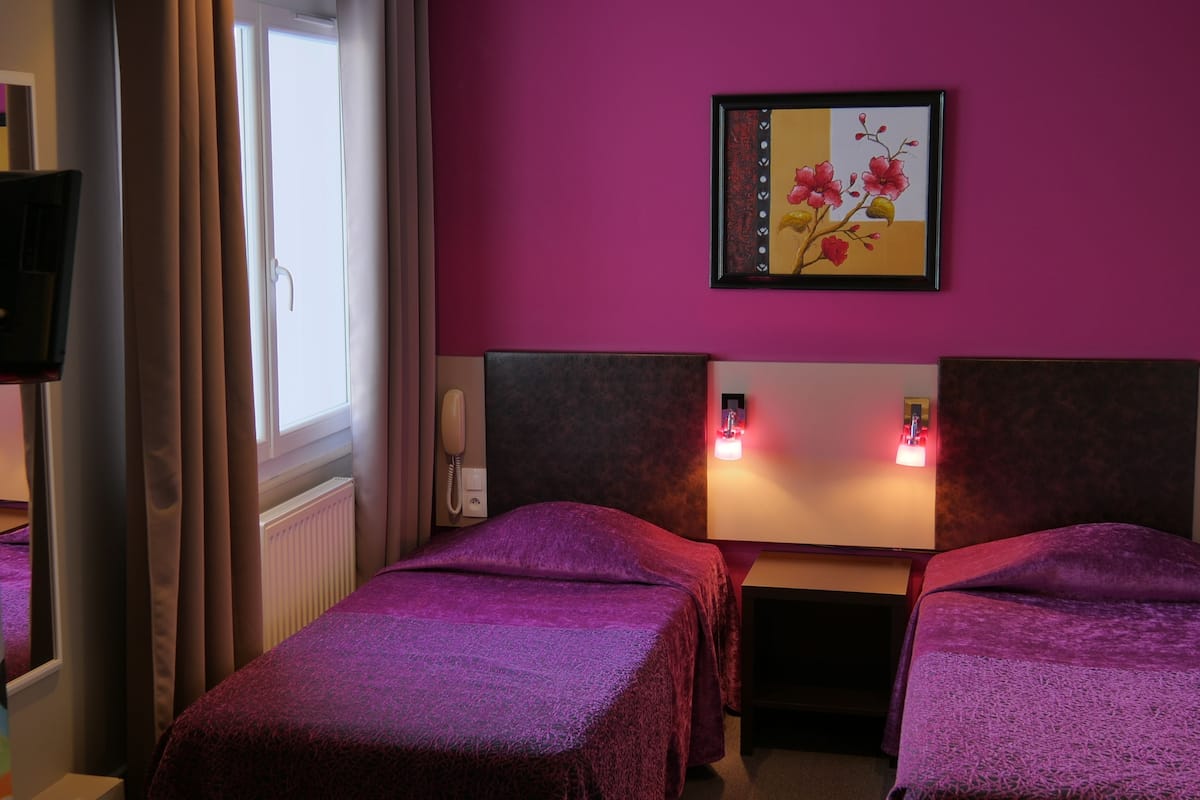 A hotel room with two beds, a window with curtains, a painting on the wall, and a bedside lamp.