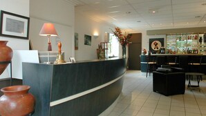 A reception area with a curved counter, a lamp, a vase with flowers, and a bar area with chairs and shelves of bottles. - Hôtel Savary (La Rochelle)