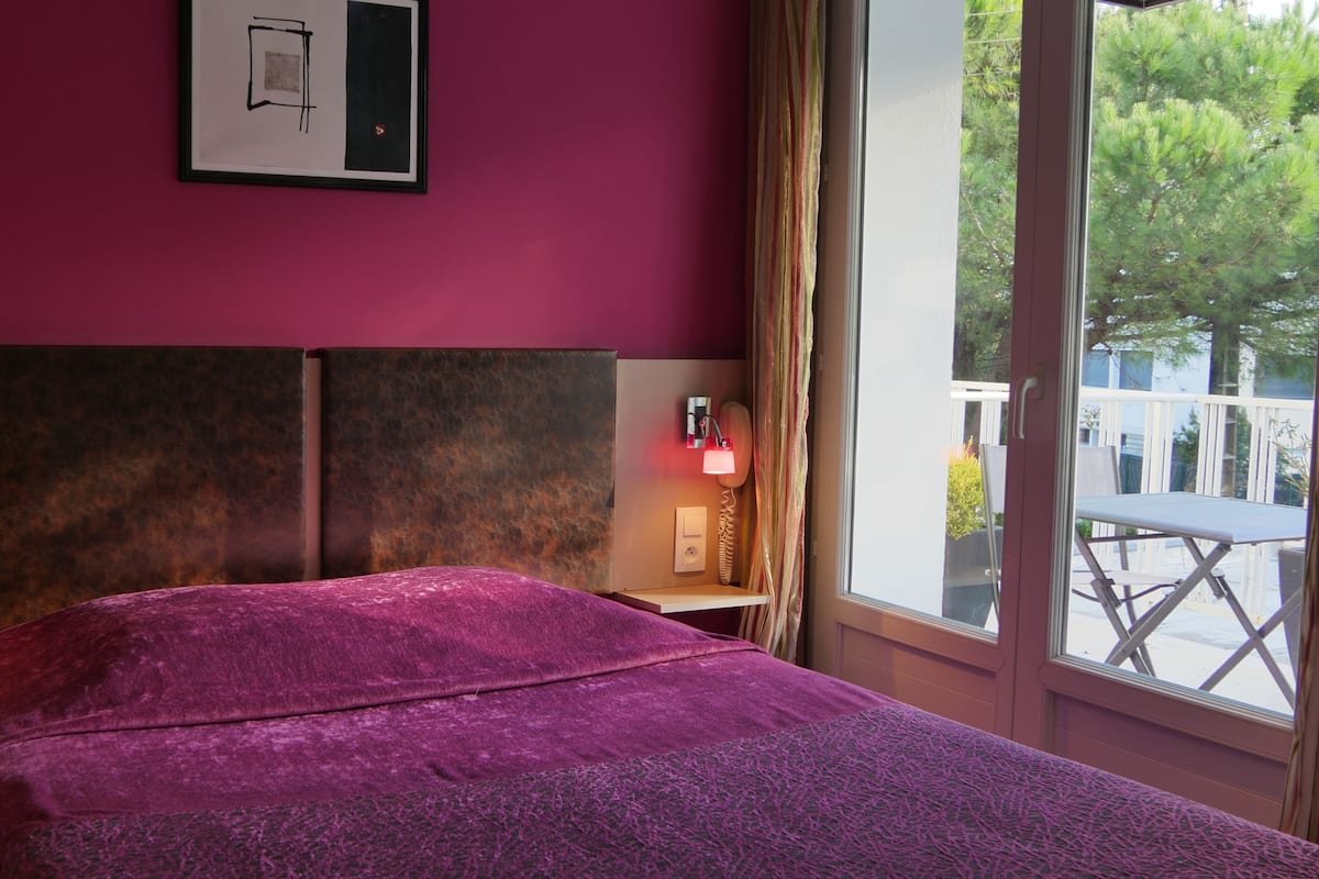 A room with a bed, two bedside lamps, a window with a view of greenery, and a wall-mounted light switch.