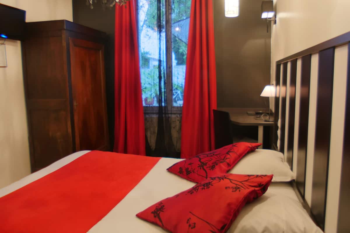 A hotel room with a bed, red curtains, a desk, and a window.